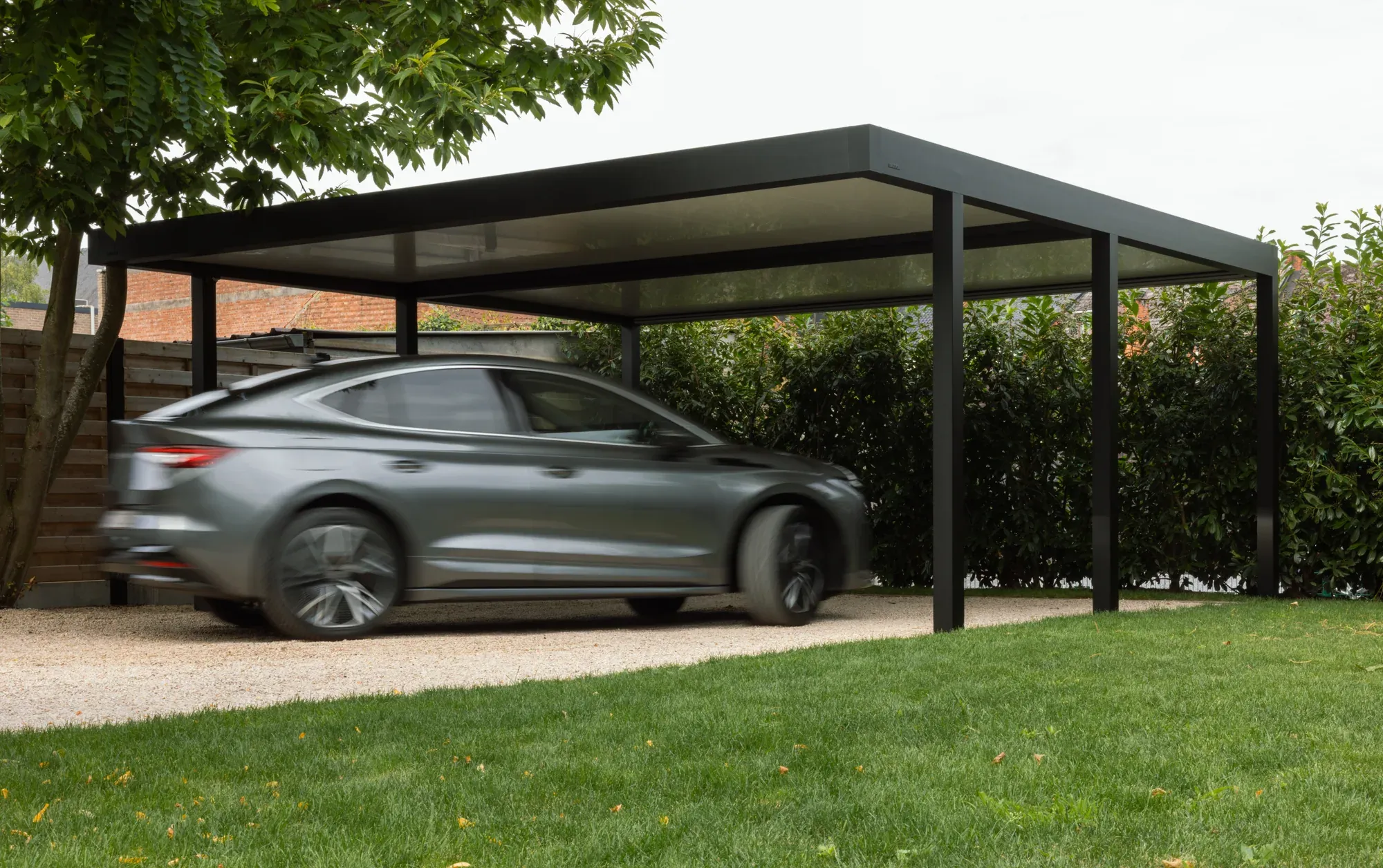 Ein Auto fährt unter einen dunklen Aluminium-Carport auf einer von Bäumen und Hecken eingefassten Einfahrt