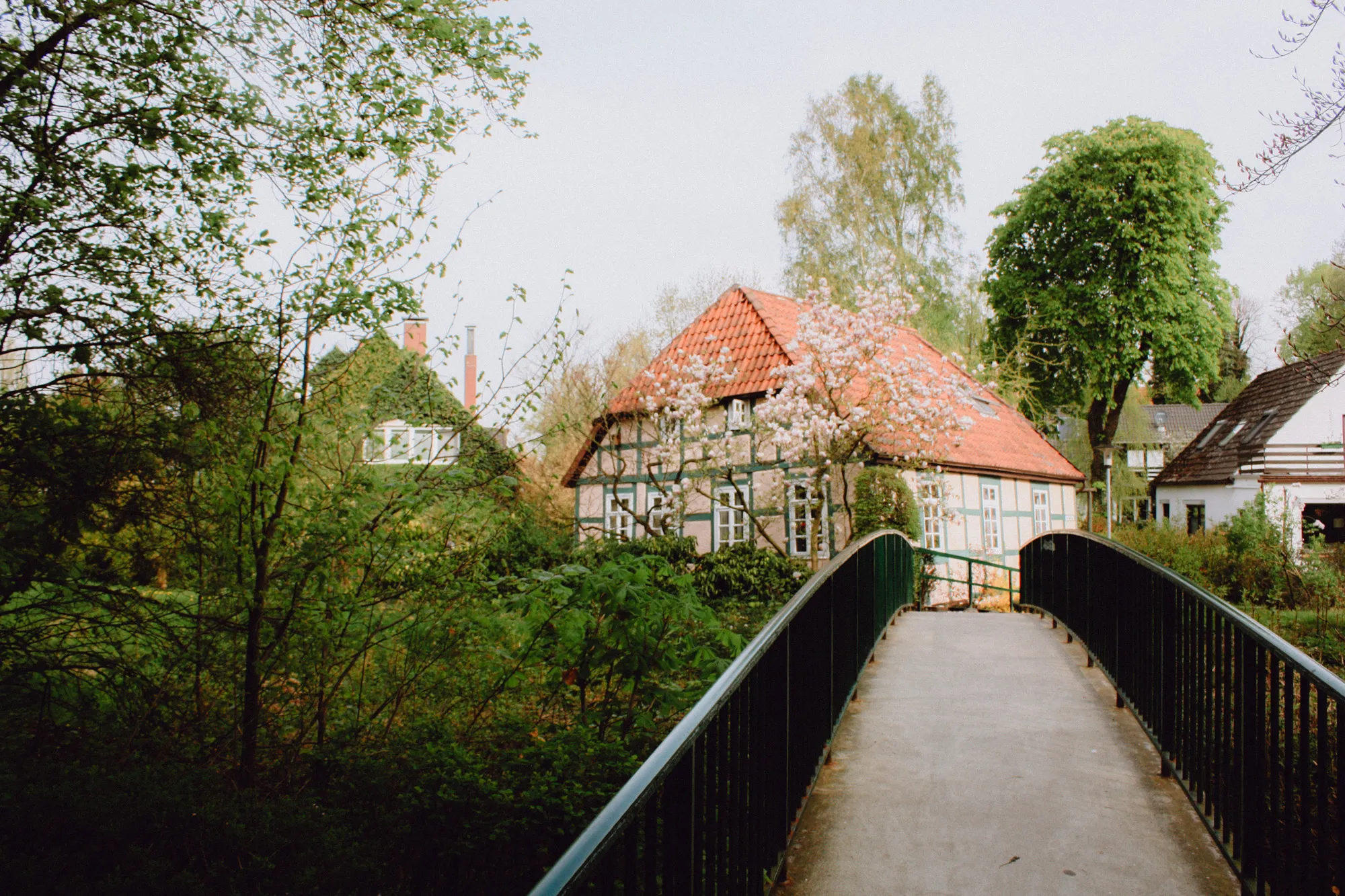 Fußbrücke vor einem Fachwerkhaus zwischen Bäumen und blühenden Sträuchern in Lilienthal